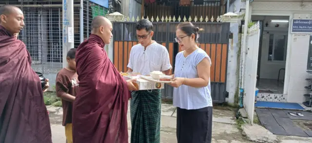 ဒေါက်တာစောနိုင်တို့ ဇနီးမောင်နှံ သံဃာအချို့ကို လှူဖွယ်လှူနေတဲ့ပုံဆိုပြီး ဒေါက်တာစောနိုင်က သူ့ ဖေ့ဘုတ်ခ်မှာ တင်ခဲ့၊ ယောဂီရောင်ဝတ် မျက်မှန်တပ်အမျိုးသမီး တစ်ဦးနဲ့ အင်္ကျီအဖြူဝတ်၊ ပုဆိုးအစိမ်းကွက် ဝတ်ထားတဲ့ မျက်မှန်တပ်အမျိုးသားတစ်ဦးတို့ သံဃာတစ်ဦးကို သင်္ကန်းနဲ့ ဆွမ်း လှူနေတဲ့ပုံ၊