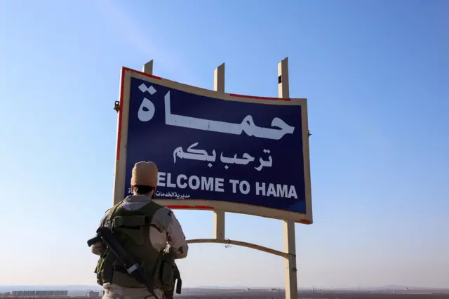 صورة لأحد مقاتلي المعارضة السورية يقف أمام لافتة عند مدخل محافظة حماة وسط سوريا.