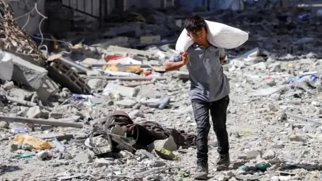 یک نوجوان پسر کیسه آرد را به دوش گذاشته و در میان ویرانه‌ها راه می‌رود