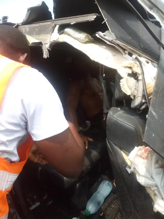 Rescue official wear white short sleeve shirt wit orange and white colour reflective jacket. E look like say e dey try tok or stretch im hand to Anthony Joshua wey sitdown inside di Lexus Jeep wey get accident.