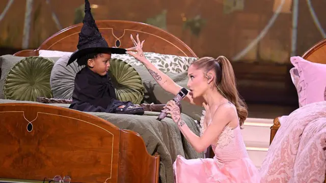 Ariana Grande cântă pentru un copil care poartă o pălărie de vrăjitoare, în timpul unei interpretări live a muzicii din Wicked.