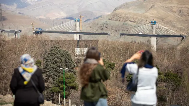 مردم پل آسیب‌دیده‌ی بی۱ را یک روز پس از تخریب آن در نتیجه یک حمله هوایی، در تاریخ ۱۴ فروردین ۱۴۰۵ در غرب تهران، در کرج، ایران مشاهده می‌کنند. مقامات ایرانی اعلام کردند که در بمباران سیزدهم فروردین این پل، هشت نفر کشته و تقریبا ۱۰۰ نفر زخمی شده‌اند. ایالات متحده و اسرائیل حمله مشترک خود به ایران را که از ۹ اسفند ۱۴۰۴ آغاز شده بود، ادامه داده‌اند. ایران در پاسخ، موجی از موشک‌ها و پهپادها را به اسرائیل و متحدان آمریکایی در منطقه شلیک کرد و در عین حال، مسیر حیاتی دریای هرمز را عملا مسدود ساخت.