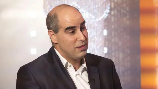 تصویری ثابت از واشی دومینگز در حال گفت‌وگو با برنامه «This Morning» شبکه ITV؛ او بار دیگر با کت‌وشلوار تیره و پیراهن سفید دیده می‌شود. پشت سر او پس‌زمینه‌ای از کریستال‌های درخشان قرار دارد که شبیه به الماس به نظر می‌رسند.