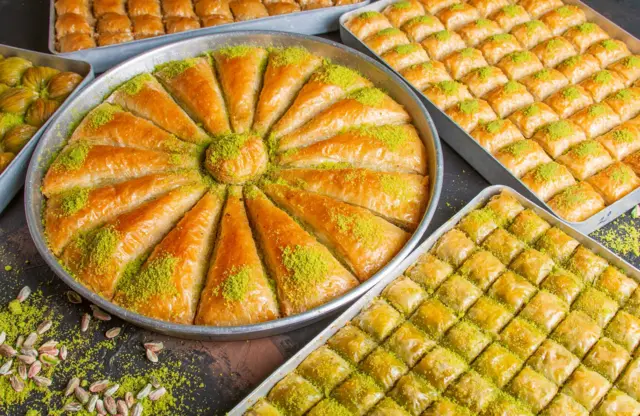 Çeşit çeşit baklavaları gösteren lezzetli bir fotoğraf. Ortadaki yuvarlak tepside havuç dilimi olarak kesilmiş üstü Antep fıstığıyla süslenmiş bir baklava yer alıyor. Etrafına yerleştirilmiş diğer tepsilerde kare ve diktörtgen kesim baklavalar mevcut. 