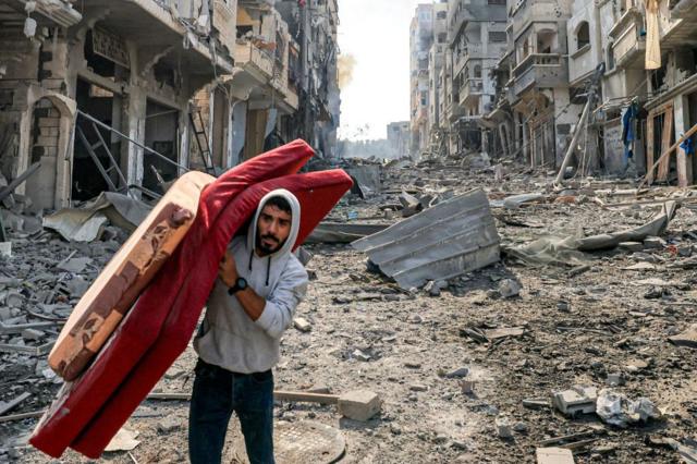 Homem caminhando com um colchão por uma rua destruída em Gaza