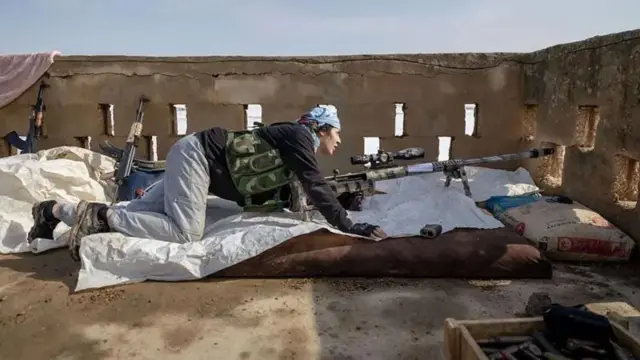 زنی کرد روی پشت بامی در کنار اسلحه تک تیرانداز نیم خیز کرده است، از روزنه لبه بام به هدفی نگاه می‌کند