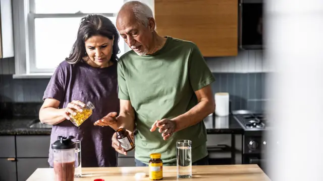 Casal na cozinha, com vários frascos de cápsulas, decidindo qual irão tomar
