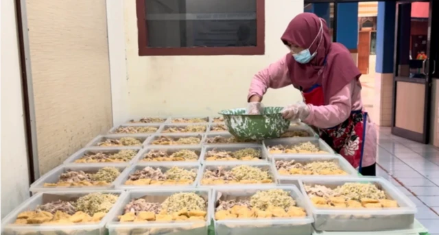 Seorang pekerja dapur sehat sedang menyiapkan suwiran ayam dan tahu goreng ke dalam nampan plastik di meja yang telah disediakan.
