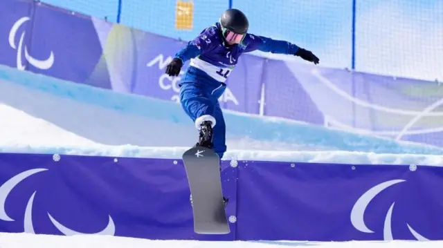 段差のあるコースでジャンプする小栗。青いウエア、黒いヘルメット、ゴーグルを着けている。前足の右脚部分には黒っぽい人工器具が見える