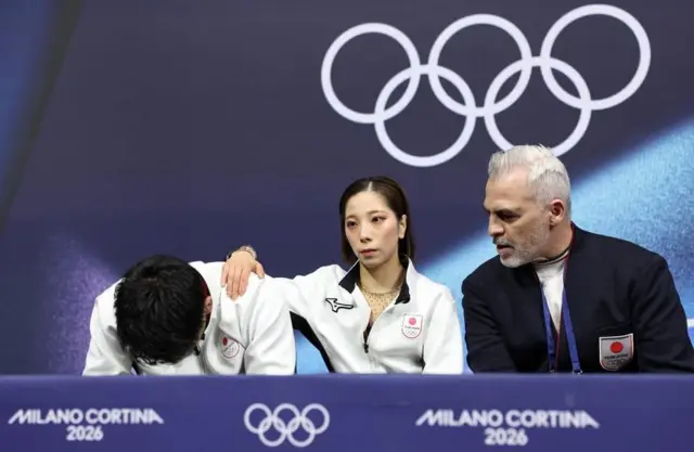 白いウエアを着た2人が横並びに座り、その横に黒っぽい上着のコーチも座っている。背後には五輪マークが見える
