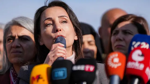 DEM Parti eş genel başkanı Tülay Hatimoğulları, çok sayıda mikrofon önünde, elinde mikrofon konuşuyor. Yanında başka partili kadınlar da var.
