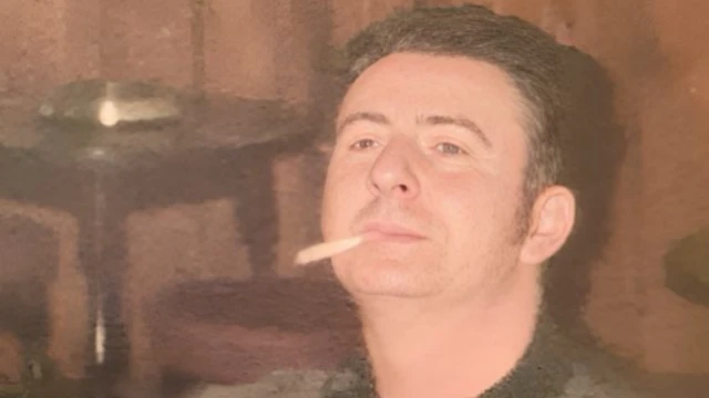 Danny está sentado em um ambiente interno que parece ser um pub, usando um suéter escuro estampado. Ele tem cabelo curto e escuro e olha levemente para cima, com um cigarro na boca