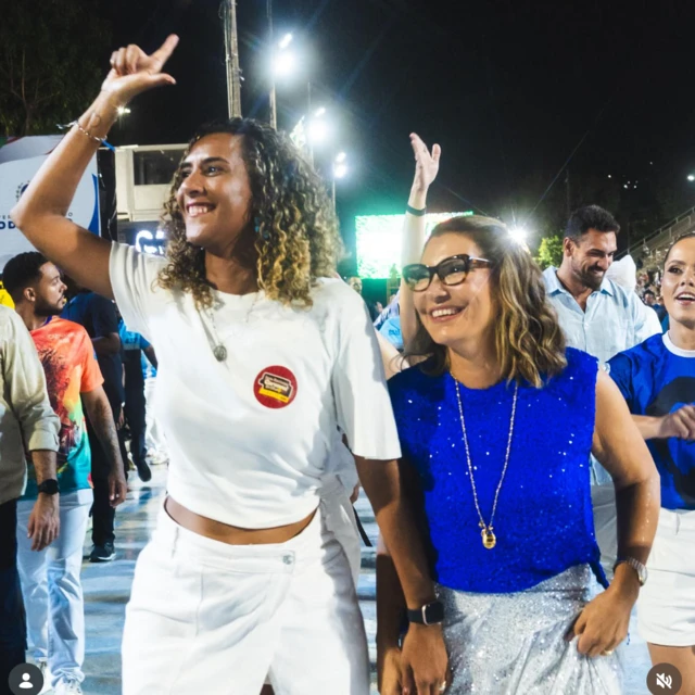 Anielle Franco vestida de branco ergue o braço direito e faz a letra L de Lula, ao lado de Janja, que veste blusa azul e saia prateada com brilho. Ambas sorriem. Ao fundo, outras pessoas participam do ensaio. 