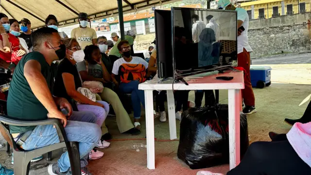 Familias intentan identificar a sus seres queridos a través de imágenes que se deslizan en dos pantallas. Algunos están sentados en sillas y con caras de agustia. 