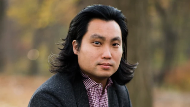 У сірому піджаку та в клітчастій сорочці, Ес Лі дивиться в камеру.