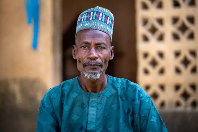 Adam Goni, wọ aṣọ olómi aro, dé fìlà, ó sì ń wo ọ̀ọ́kán