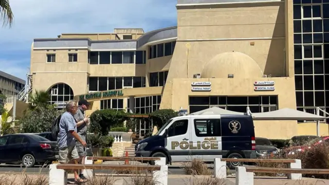 An Egyptian police van standing outside a hospital with two male tourists walking by. රෝහල ඉදිරිපිට ඊජිප්තු පොලිස් වෑන් රථයක් නවතා ඇති අයුරු සහ ඒ ඉදිරියෙන් සංචාරකයින් දෙදෙනෙකු ඇවිද ගෙන යන ආකාරය.