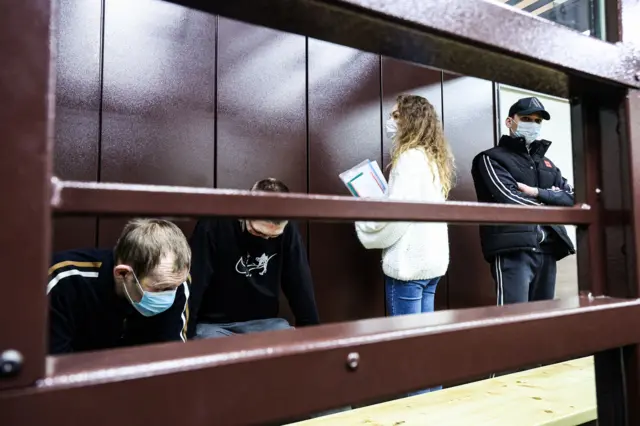 Коричневая клетка в зале судебных заседаний. В клетке находятся четыре человека в медицинских масках. Слева направо: сидящий мужчина в спортивной кофте, сидящий мужчина в черной кофте и серых штанах. Третьей спиной к камере стоит женщина с длинными волосами, в белом свитере и синих джинсах. В руках женщина держит папки с бумагами. Последним стоит мужчина, скрестив руки на груди. Он одет в спортивную одежду, на голове бейсболка