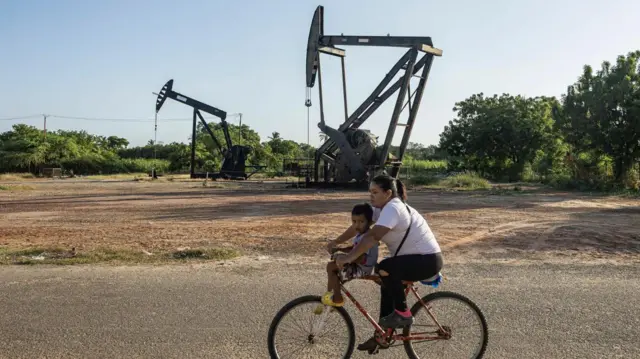 A woman rides a bicycle with a child in front of oil derricks in Cabimas, Venezuela, 18 December 2025. US President Donald Trump announced a total blockade of all sanctioned oil tankers entering and leaving the country, a strategy analysts say is intended to be selective and part of growing pressure over crude oil and other natural resources.