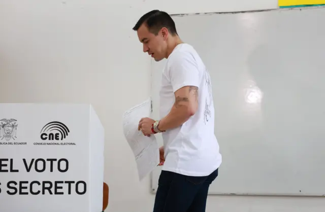 Daniel Noboa, con pantalón negro, camiseta blanca y brazos tatuados, con un papel en la mano en un centro de votación. 