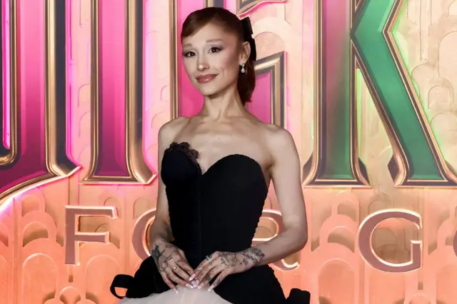 Ariana Grande viste un vestido blanco y negro y se encuentra frente a una valla publicitaria que promociona la película Wicked: For Good.