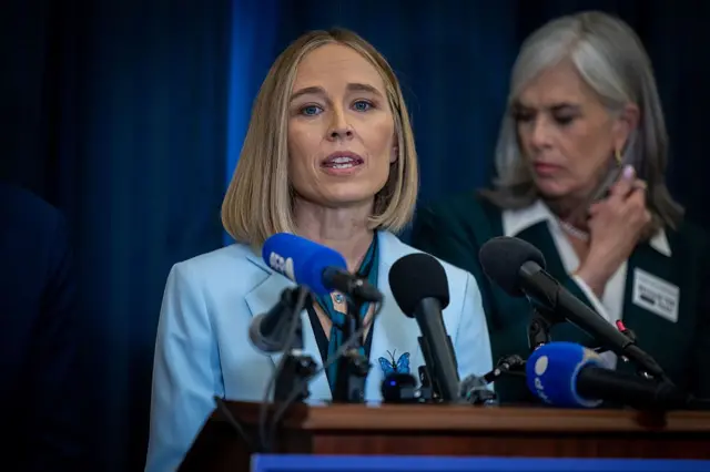 Açık mavi takım elbiseli sarışın bir kadın mikrofon başında. Arkada beyaz saçlı başka bir kadın gözüküyor.