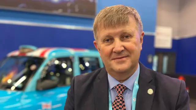 Un hombre con traje y corbata a cuadros con la bandera británica. Un taxi londinense azul claro está detrás de él, desenfocado.