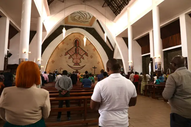 Worshippers dey stand during mass inside one large Catholic church, Our Lady of Victoties Cathedral for Yaounde featuring high ceilings and large pillars and wall murals.