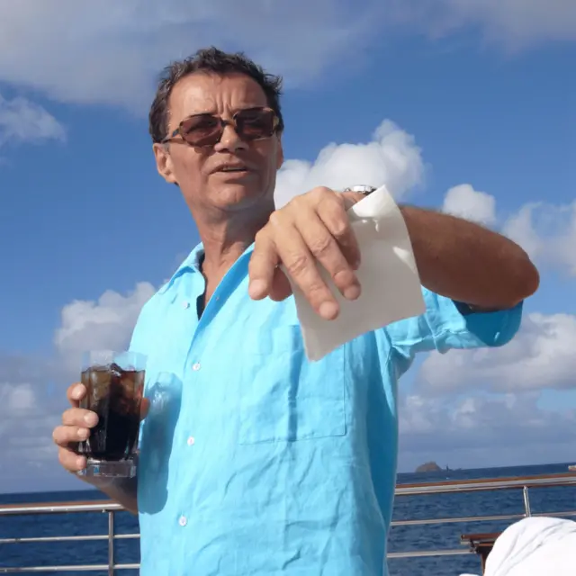 一名男子身穿浅蓝色短袖衬衫，戴着太阳眼镜，一手拿着饮料，一手拿着一张纸。他留着深色短发。背景中可以看到广阔的水面和蓝天白云。