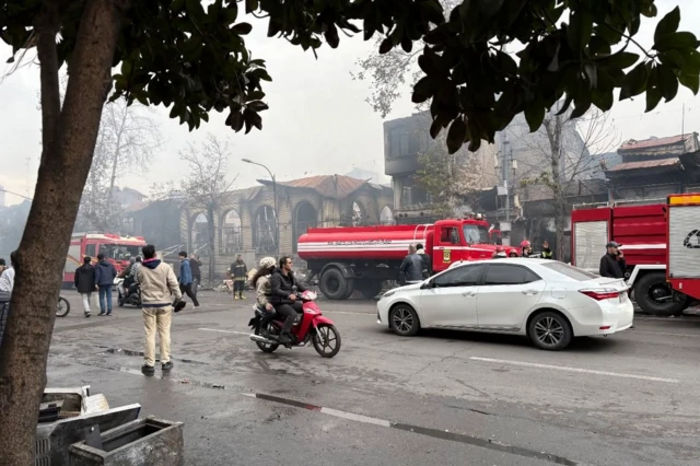 خیابانی در حاشیه بازار رشت در حالی که چند ماشین آتش‌نشانی در تصویر دیده می‌شوند. در تصویر دود سفیدی دیده می‌شود. چند نفر از عابران به ناحیه سوخته‌شده نگاه می‌کنند.