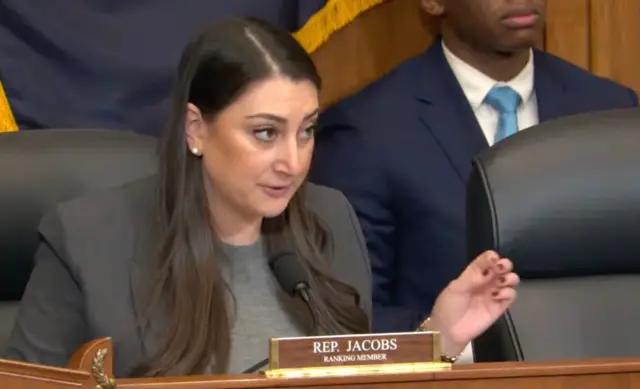 Sara Jacobs uri mu bahagarariye leta ya California mu nteko y'Amerika, yicaye arimo kuvugira mu ndangururamajwi, akoze ikimenyetso n'ikiganza. Imbere ye hari ikirango cyanditseho izina rye.