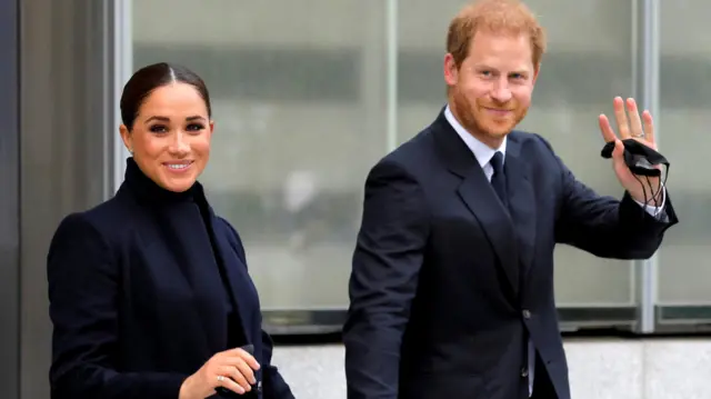 Meghan e Harry sorrindo para foto enquanto caminham com trajes formaiswazamba como funcionaárea externa