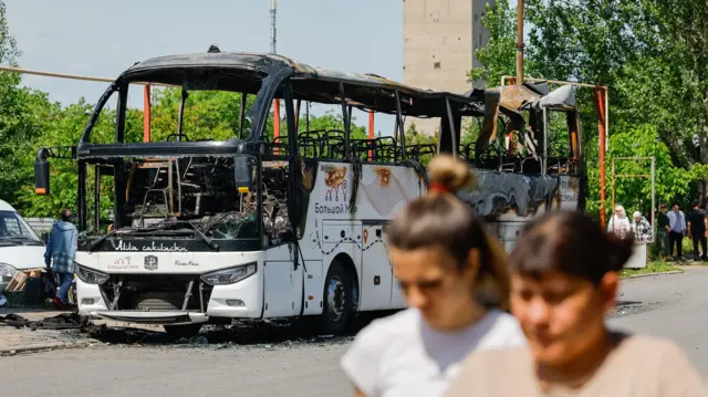 Две женщины идут по улице, за ними — сгоревший автобус.