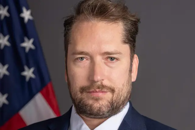 Retrato de Darren Beattie, que aparece sério em frente a bandeira dos EUA