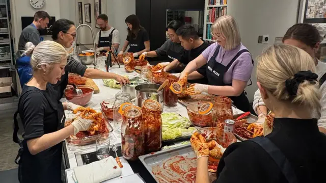 10 человек стоят у длинного стола и втирают красную пасту в кусочки капусты