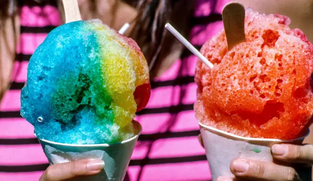 Girl holds two rainbow coloured slushies in giant ice cream cones.