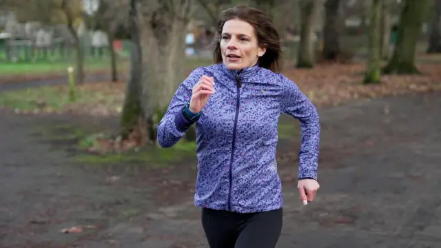 Ruth Clegg, uma mulher de cabelos castanhos e jaqueta esportiva roxa, corre em uma pista que atravessa um parque.