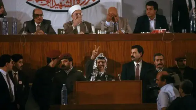 Kiongozi wa PLO Yasser Arafat akionyesha ishara ya ushindi katika kikao cha Baraza la Kitaifa la Palestina huko Algiers mnamo 1988.