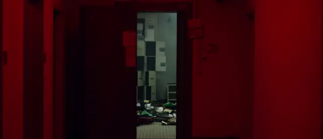Pasillo iluminado en rojo con una puerta abierta que muestra una sala con casilleros y papeles esparcidos en el suelo