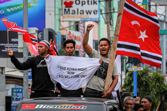 Warga mengibarkan bendera Gerakan Aceh Merdeka (GAM) sembari mengawal bantuan kemanusiaan yang ditujukan bagi para penyintas banjir dahsyat yang baru saja melanda Sumatra, di Bireuen, Provinsi Aceh, pada 25 Desember 2025. Meskipun pemerintah Indonesia telah berupaya, rasa frustrasi kian memuncak akibat lambatnya penanganan bantuan; hal ini memicu desakan kepada pemerintah pusat agar menetapkan status bencana nasional dan mengizinkan masuknya bantuan internasional ke wilayah-wilayah terdampak. Pemerintah Provinsi Aceh menyatakan telah meminta bantuan kepada sejumlah badan Perserikatan Bangsa-Bangsa (PBB), dengan merujuk pada pengalaman kampanye bantuan badan-badan tersebut pascatsunami tahun 2004. (Foto oleh