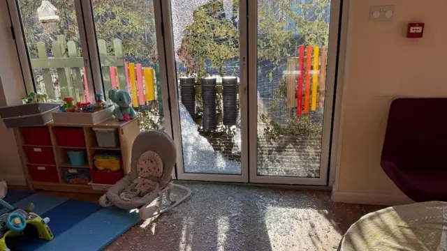 Se muestra un área de juegos infantiles con muchos fragmentos de vidrio esparcidos por todas partes. La puerta de cristal está rota.