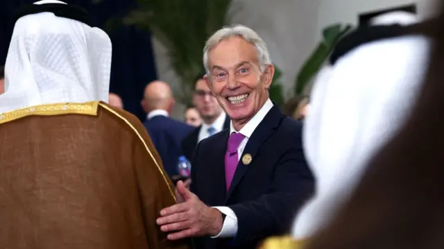 El ex primer ministro británico Tony Blair durante la cumbre de Sharm El-Sheij, Egipto, el 13 de octubre de 2025. Blair sonríe y coloca su mano en el brazo de un hombre que viste la tradicional ropa árabe. Otro hombre de vestimenta similar está en la imagen. 