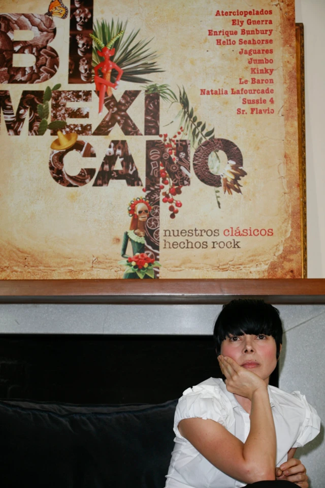 Ely Guerra, con una camisa blanca, está sentada con la mano en la barbilla. Detrás tiene la portada del álbum que dice “Bimexicano, Nuestros Clásicos Hechos Rock”.