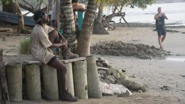 Una persona negra contempla el atardecer sentado sobre un muro de contención frente al mar mientras algunos turistas recorren la playa.