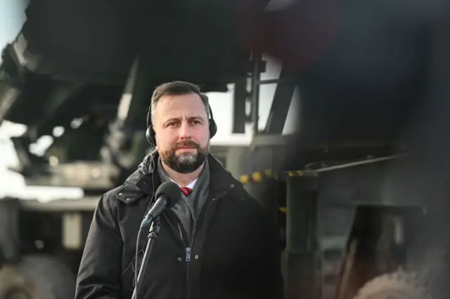 Косіняк-Камиш у зимовій куртці та навушниках із кабелем виступає на брифінгу. На задньому плані видно пускові установки комплексу ППО Patriot.