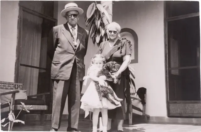 Alberto Veraza, vestido de traje, con corbata y sombrero, tiene las manos hacia atrás. A su lado está Adriana, quien tiene el cabello blanco, lleva lentes y un vestido oscuro. Muy cerca de ella está Mara, que tiene trenzas y un vestido blanco y carga un perro. Los tres miran a la cámara y están en lo que parece ser el patio de una casa