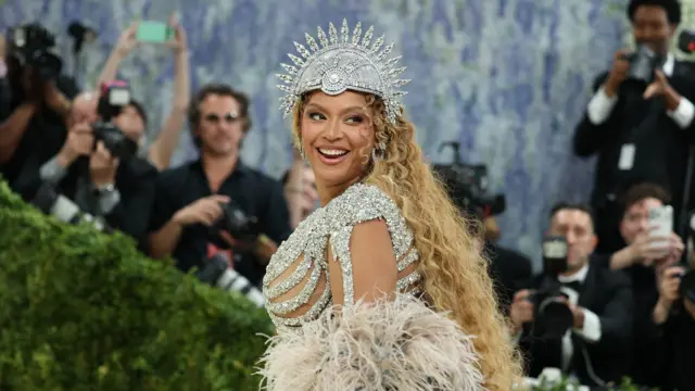 Zbliżenie na kreację Beyoncé, która ma na sobie bogato zdobioną, jasną suknię z motywem szkieletu oraz długą, obszerną pelerynę z szarego futra. Na głowie widoczna jest ozdobna, promienista korona z metalicznymi elementami. W tle znajdują się fotografowie z aparatami.