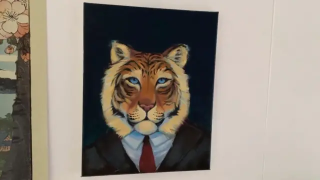 A picture hanging on a white wall. It shows a tiger's head, with striped fur and piercing blue eyes, sitting on top of a dark suit, white shirt and red tie.