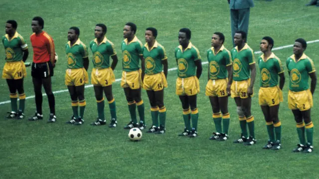 L'équipe de football du Zaïre alignée pour une photo d'équipe avant un match de la Coupe du monde 1974. Les joueurs portent des chemises vertes à l'effigie d'un léopard sur la poitrine, des shorts jaunes, des chaussettes vertes et des bottes noires. Le gardien de but porte un maillot rouge à manches longues, un short noir et des chaussettes noires