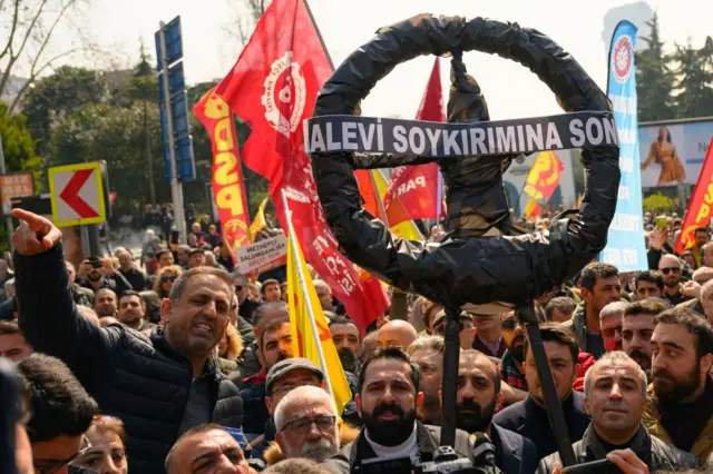 9 Mart'ta İstanbul'da toplanan Alevi kurumlarının basın açıklamasından bir fotoğraf. Grup içinde farklı bayraklar ve üzerinde "Alevi soykırımına son" yazan 
siyah bir çelenk var. 